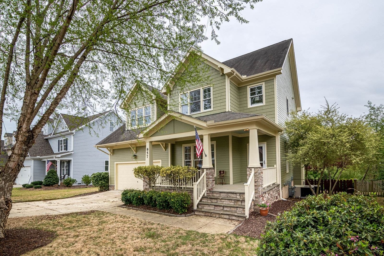 Two-story beige house with front porch, white railings, brick foundation, and American flag
